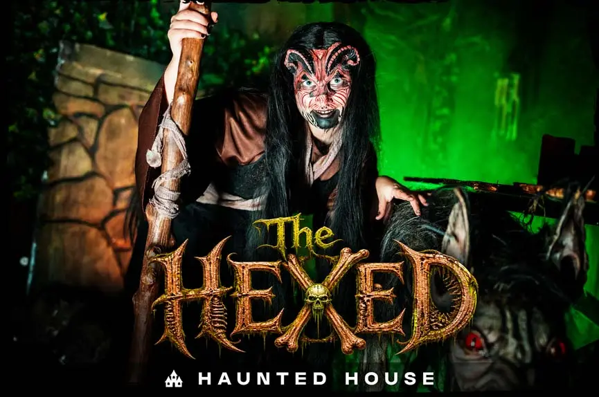 A demonic witch with red-striped horns and dark hair crouches with a wooden staff in eerie green lighting to promote The Hexed Haunted House.